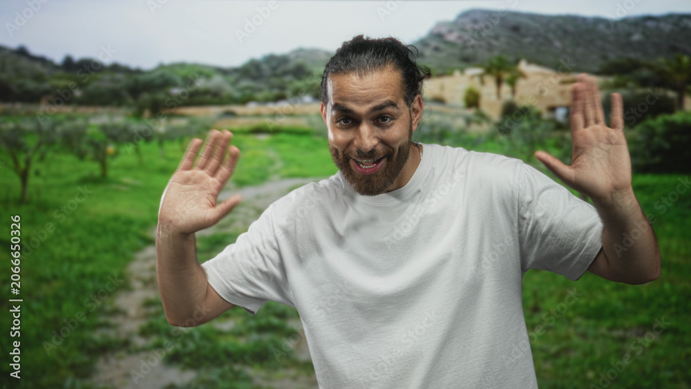 Naklejka premium Man raising hands with palms visible and beard, wearing white shirt and necklace, standing on a country path in forest, smiling; playful greeting.