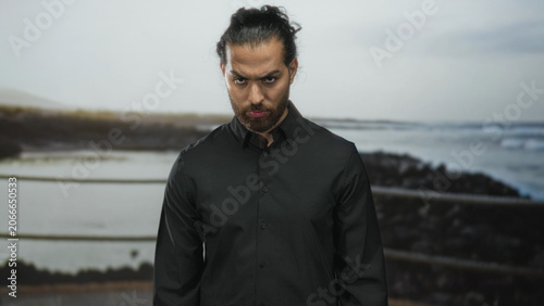 Tableau sur toile Man, young arab hispanic with beard and tied hair, in black shirt furrows brow and looks down at seaside railing on rocky coast; brooding solitude