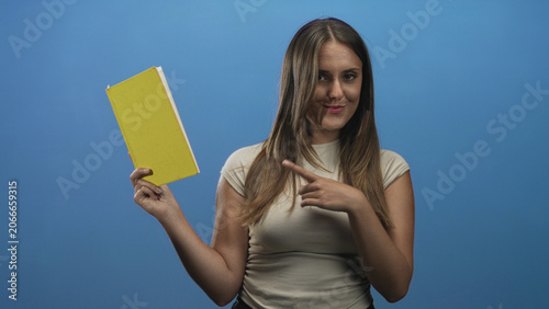 Картината върху платно Woman holding a yellow book points finger to the cover in a studio, smiling while presenting the hardcover; confidence learning inspiration