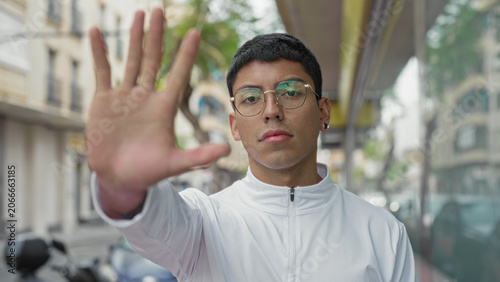 Fotografie Man holds palm to camera in a stop gesture on street, wearing white jacket and glasses; defiance privacy