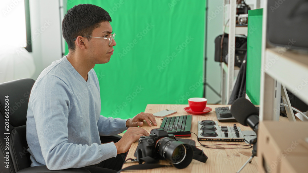 Fototapeta premium Man photographer with glasses types on keyboard, then lifts hands to head beside camera, control dial and red cup in studio with green screen; creative frustration.