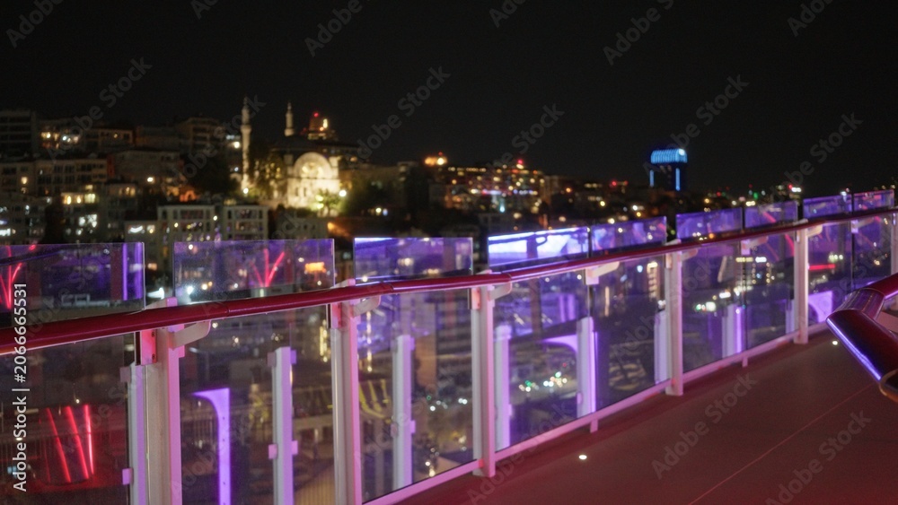 Fototapeta premium Night rooftop terrace glass balustrade in sharp foreground with blurred urban skyline and colorful bokeh lights, defocused night view city; background backdrop copyspace calm.