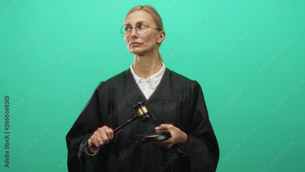 Naklejka premium Woman judge in black robe raises gavel with right hand over wooden sounding block in studio green screen, wearing glasses and white collar; authority justice.