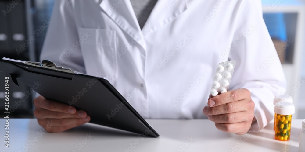 Fototapeta premium Pharmacist holding blister with pills and clipboard at white table in clinic, closeup