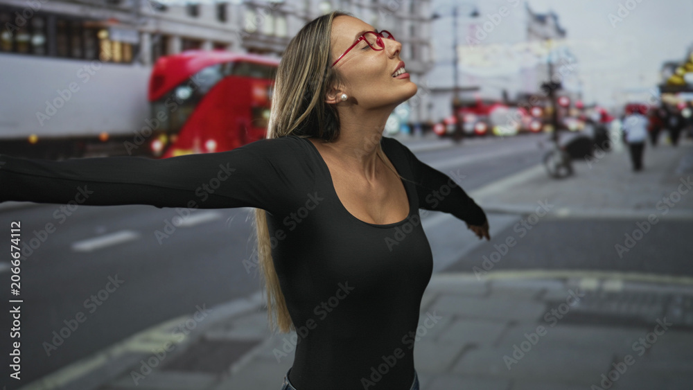 Fototapeta premium Young woman smiling with arms outstretched on a busy city street beside red double decker bus, looking upward at the sky; joy exploration.
