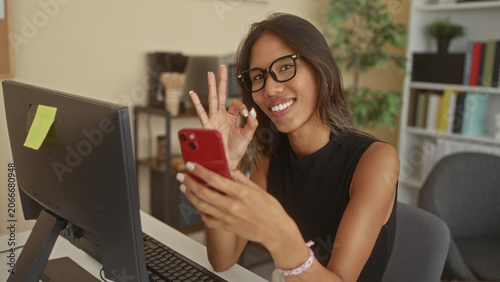 Canvas Print Thai woman with glasses holding red smartphone and making ok sign while seated at desk with monitor and keyboard in office building; remote work joy