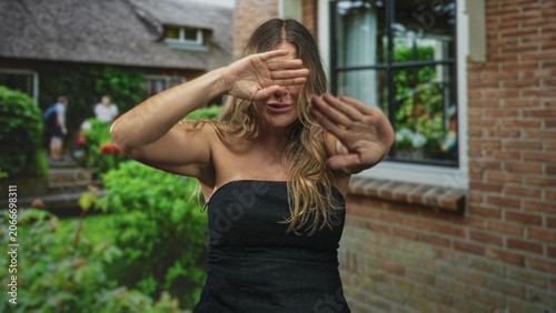 Foto Woman shields eyes with hands and holds palms forward in front of brick building window and garden; shyness privacy solitude