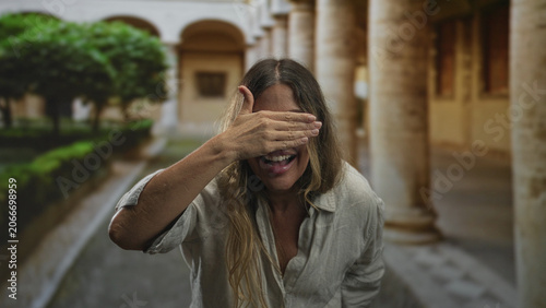 Obraz na płótnie Woman covers eyes with hands and laughs inside a building courtyard framed by stone columns and trimmed hedges; joy playfulness