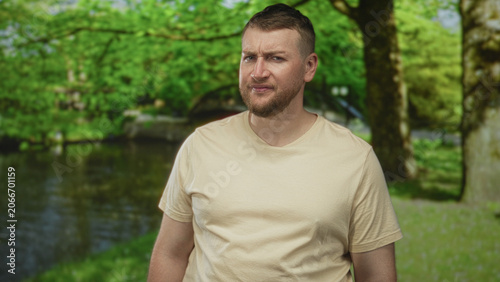 Obraz na plátně Man grimacing with visible face and stubble, wearing beige tshirt, standing in forest by a pond and small bridge among trees and grass; disgust