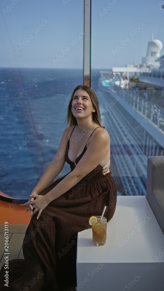 Obraz premium Woman smiling and sitting showing cleavage on cruise ship terrace with ocean view and drink nearby; relaxed serenity.
