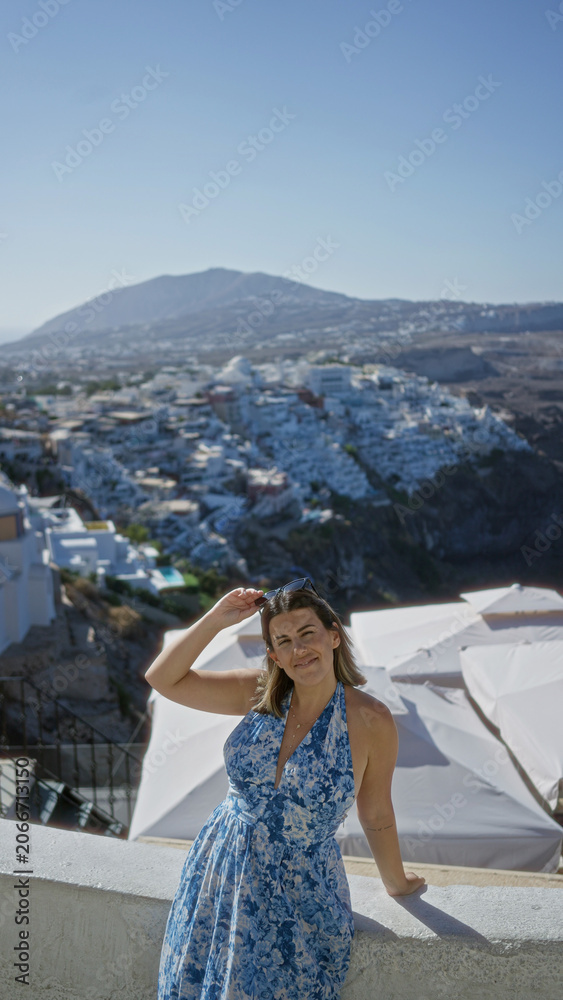Fototapeta premium Woman raising arm with sunglasses on a white building terrace, leaning on a wall and smiling at the view; vacation serenity.