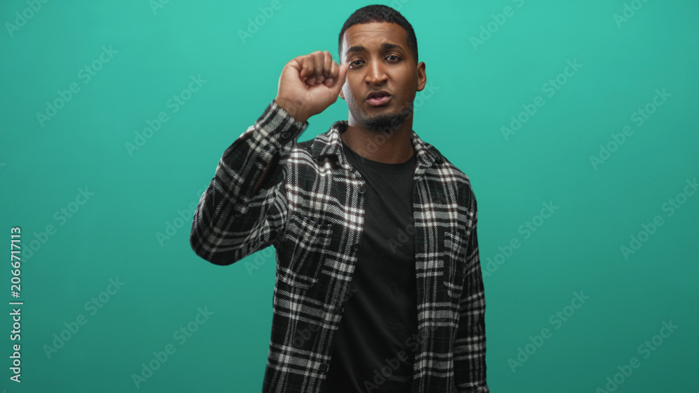 Naklejka premium Young black man holding electric screwdriver in right hand while speaking and gesturing toward camera in a studio with teal backdrop; practical confidence.