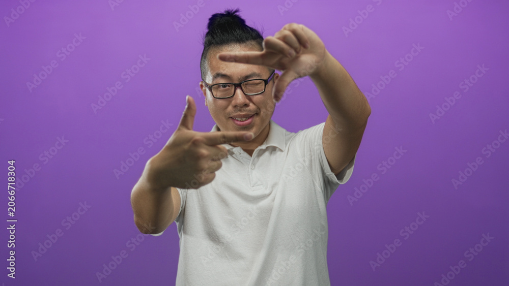 Fototapeta premium Man framing with hands and winking eye in purple studio set wearing white polo and glasses with top knot; playful creativity.