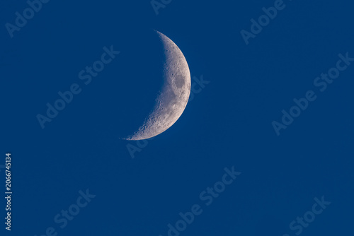 Close up of a waxing crescent moon with detailed craters against a deep blue sky background.