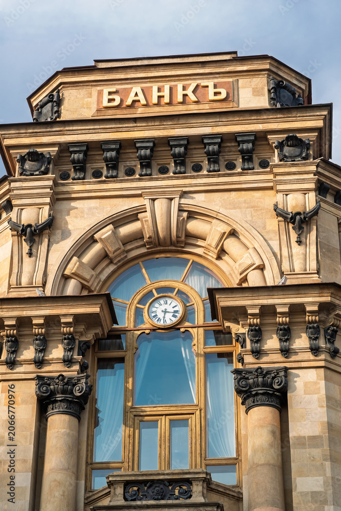 Naklejka premium Moscow, Russia - May 21, 2026: Historic bank building showcasing intricate stone architecture, large windows, and a clock, with the word BANK prominently displayed