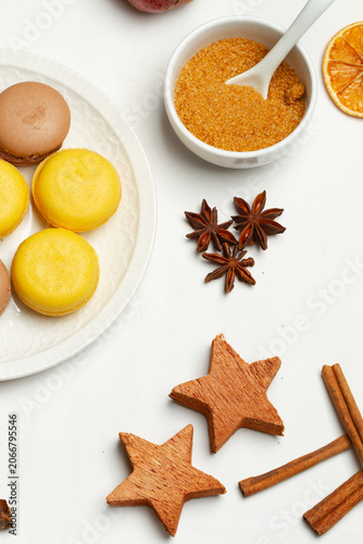 Unos macarons junto a unas ramitas de canela, azucar morena, y anis estrellado sobre un fondo blanco. Vista superior y de cerca.