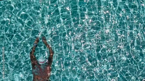 Aerial overhead view of a man swimming underwater in a clear turquoise swimming pool with bright shimmering sun reflections