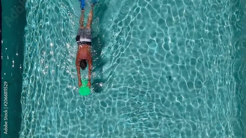 Aerial top down view of a male swimmer training in a clear blue swimming pool with a kickboard and swim fins