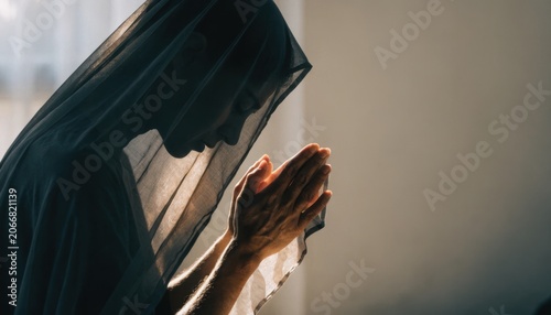 Woman wearing a dark veil is praying with closed eyes and hands clasped, captured in a serene and contemplative moment with soft lighting.