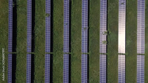 Aerial view captures solar panels placed in organized rows on a grassy area. Sunlight reflects off the panels while some trees are visible nearby