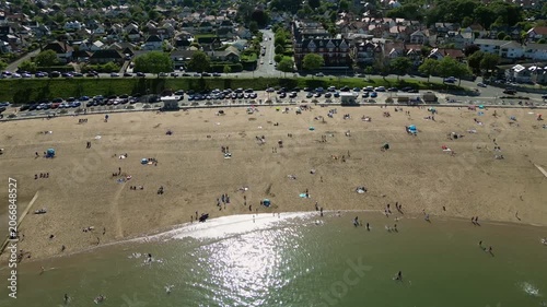 People are on Colwyn Bay Beach in North Wales. Some walk along the shore while others sunbathe or swim in the water. Cars are parked nearby and houses are visible in the background