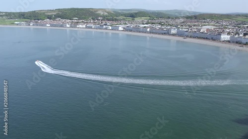 A boat moves through the ocean water near Llandudno in North Wales. The camera shows the coastline and buildings along the shore on a clear day