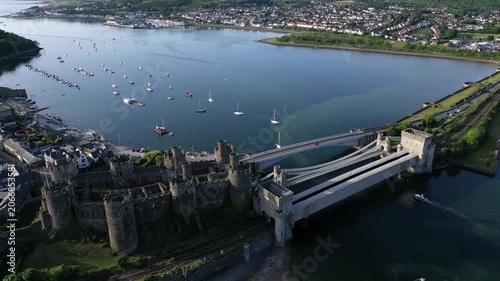 Conwy Castle stands tall near the water as boats sail in the background. People enjoy the view and walk along the shore. Green hills surround the area
