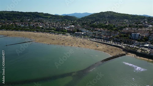 Colwyn Bay Beach in North Wales on a sunny day. People explore the beach, swim in the sea, and relax on the sand. The view highlights the shoreline and surrounding area