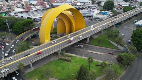 Aerial drone footage showcases Guadalajara's iconic Millennium Arches monument with heavy highway traffic navigating the urban landscape.