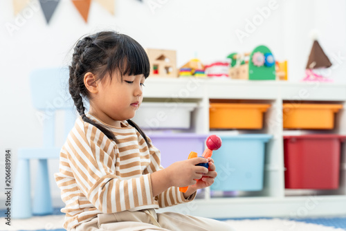 Foto Asian toddler girl sits on foam mat in playroom, grips colorful toy with calm at