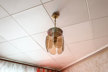 vintage-style pendant lamp with amber glass petals hangs from a white Armstrong ceiling; floral wallpaper and sheer curtains frame the corner