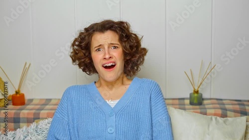 young woman with curly hair and braces reacts with wide eyes and an open mouth, expressing intense shock and fear while sitting comfortably at home