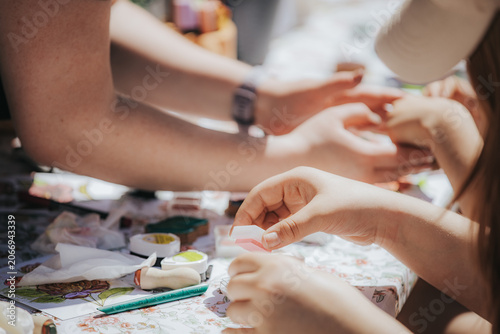 Hands of children and adults engaged in arts and crafts activity at a table covered with colorful materials and tools under bright sunlight outdoors