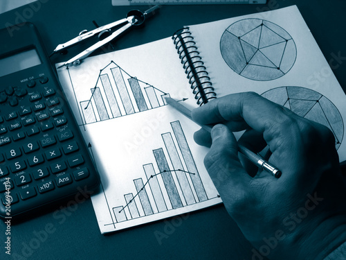 Hand of a person pointing with a pencil to colorful business charts and graphs in a notebook on a red desk