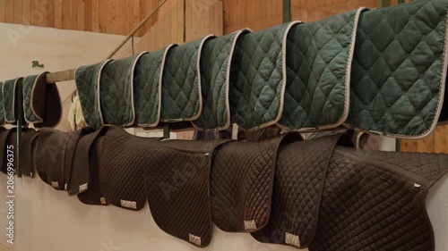 Equestrian saddle pads hanging on storage racks inside a professional stable. Rows of quilted horse blankets and riding equipment for equestrian sports training on a countryside ranch farm.