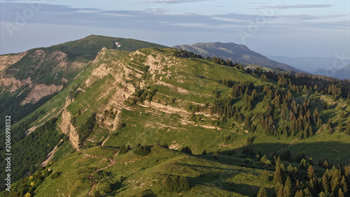 Aerial view of Col de la Faucille mountain pass with lush green slopes, rocky ridges, and coniferous forests under soft morning light Gex, Auvergne-Rhone-Alpes, France.