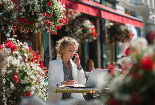 A woman in a white blazer sitting at an outdoor cafe table, talking on her mobile phone with a laptop computer open in front of her, flowers