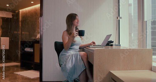 Young woman in blue dress drinking coffee while working on laptop at desk in luxury hotel room. Modern interior with city view window and marble bathroom background. Side view medium shot.