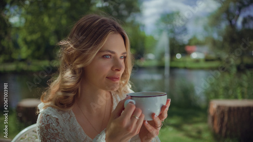 Woman sitting by a table in an outdoor cafe and holding a white cup and enjoying the scent