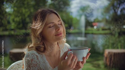 Woman sitting by a table in an outdoor cafe and holding a cappuccino cup and enjoying the scent