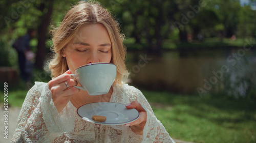 Woman sitting by a table at a park in an outdoor cafe and holding a white cappuccino cup and drinking