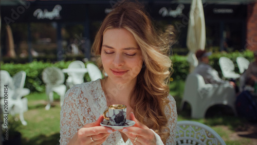 Woman sitting by a table in an outdoor cafe and holding a tiny elegant espresso cup up to the camera