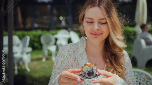Woman sitting by a table in an outdoor cafe and holding a tiny elegant espresso cup up to the camera