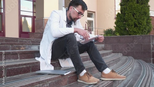 Young doctor reading medical report while sitting on stairs outside thospital. Tired medical cardiologist examines electrocardiogram of patient in front of hospital. Healthcare and medicine.