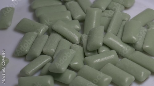Close-up of a pile of green chewing gum pellets rotating on a white plate, showcasing their smooth glossy coating under bright studio lighting.
