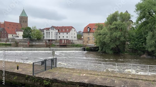Scenic view of the Ems river weir in Rheine, Germany, with historic buildings, a church tower, and green trees lining the waterfront under an overcast sky.