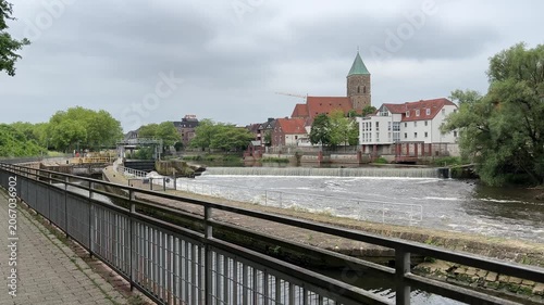 Scenic view of the Ems river weir in Rheine, Germany, with historic buildings, a church tower, and green trees lining the waterfront under an overcast sky.