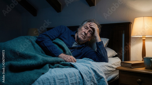 Worried Senior With Hand On His Head Lying in a Bedroom Under Teal Blanket