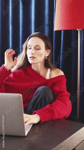 Concentrated young woman with wireless earbuds typing on laptop at home, focused girl working remotely, tired female freelancer fixing hair while studying in cozy room with red lamp, vertical