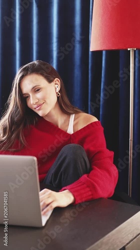Young woman in a red sweater, with wireless earbuds, typing on a laptop, smiling at the screen, sits at a desk with an angled leg against dark blue curtains and a red floor lamp, home interior